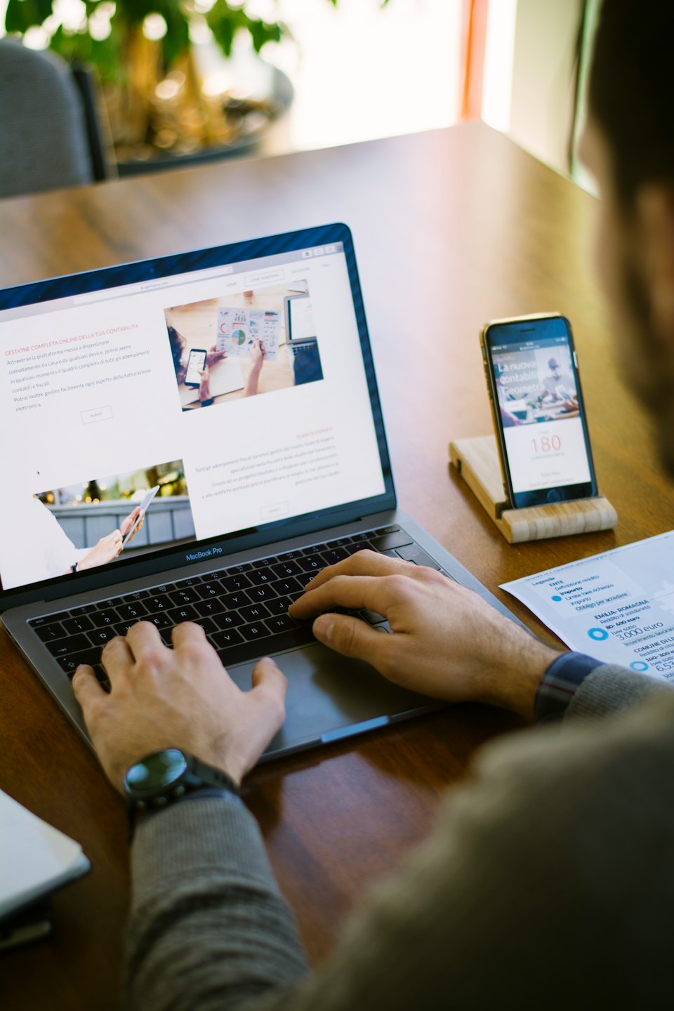 Persona che digita su un MacBook con iPhone e documenti sulla scrivania.